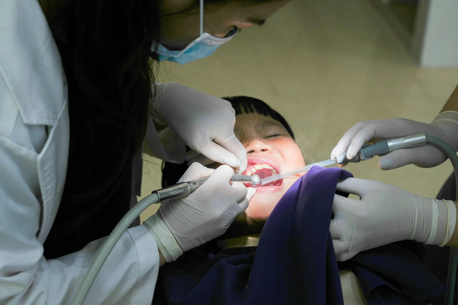 A young child receiving dental treatment from a masked dentist using dental tools indoors.